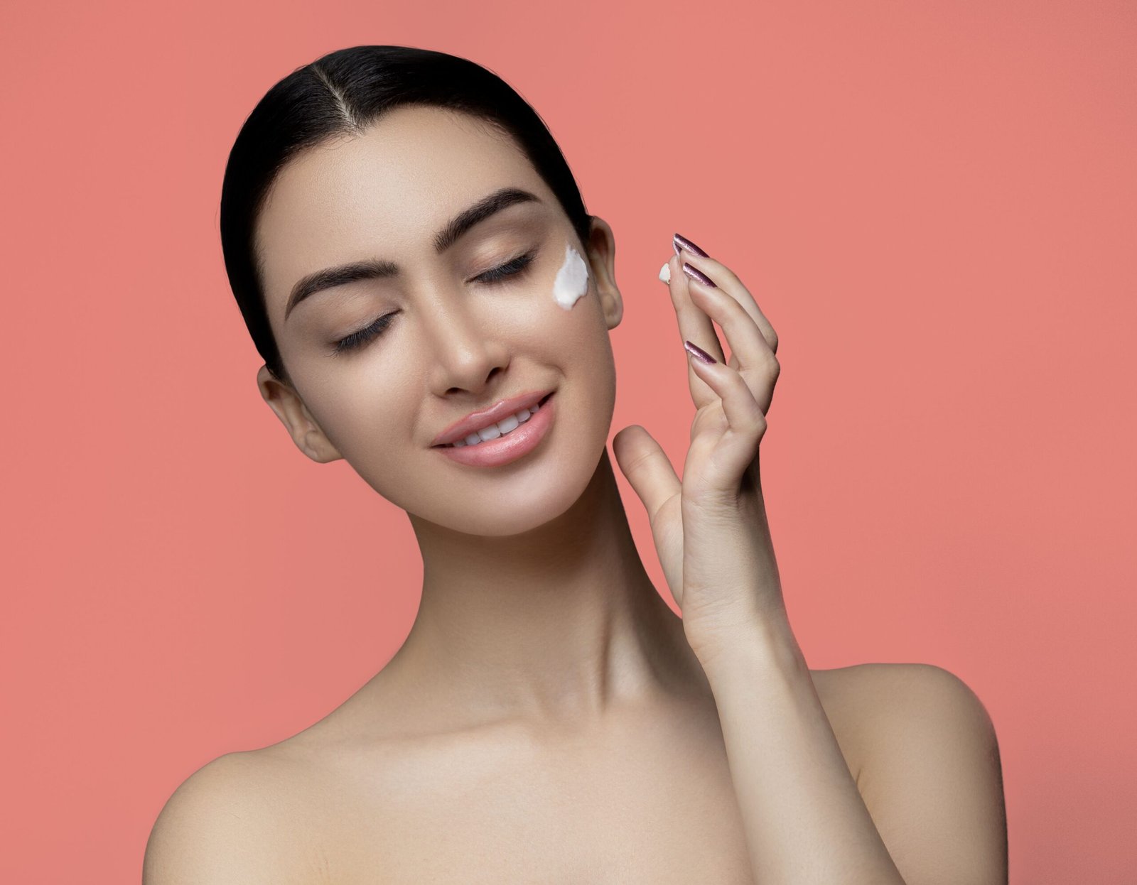 Studio portrait of a smiling woman applying cream on her face against a pink background.