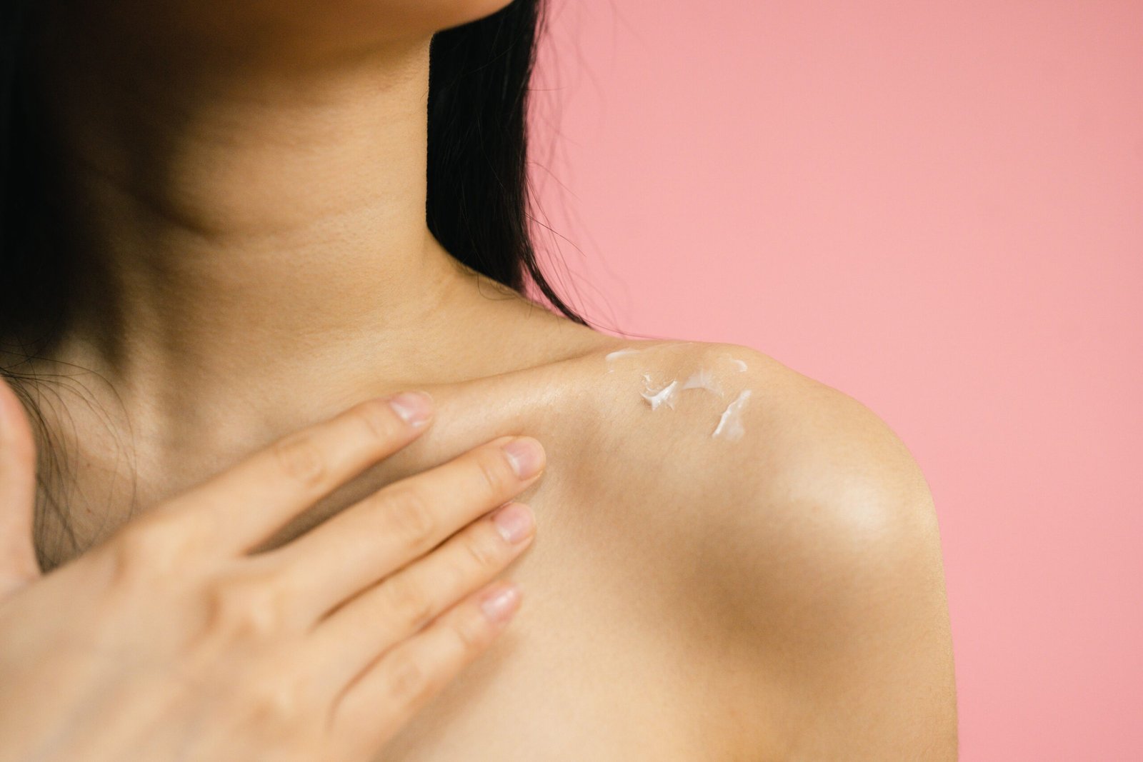 A woman applies a moisturizing cream to her shoulder against a pink background.